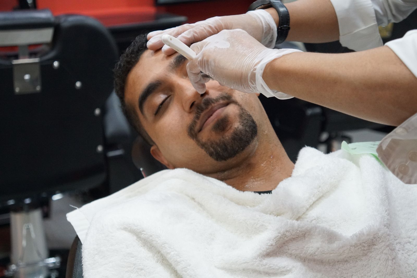 A client relaxing with a towel across his chest while a barber in gloves shapes his brow