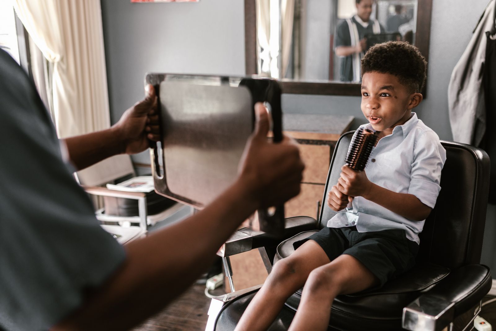 A young boy holding a brush like a microphone while the barber holds up a mirror
