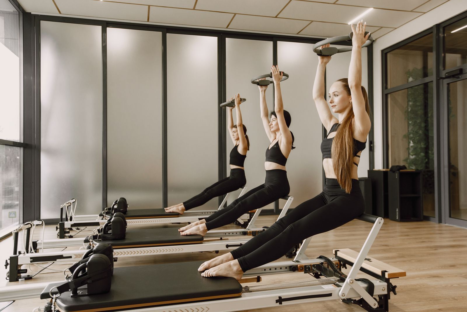 Three people on reformers in a bright studio, arms raised holding Pilates rings