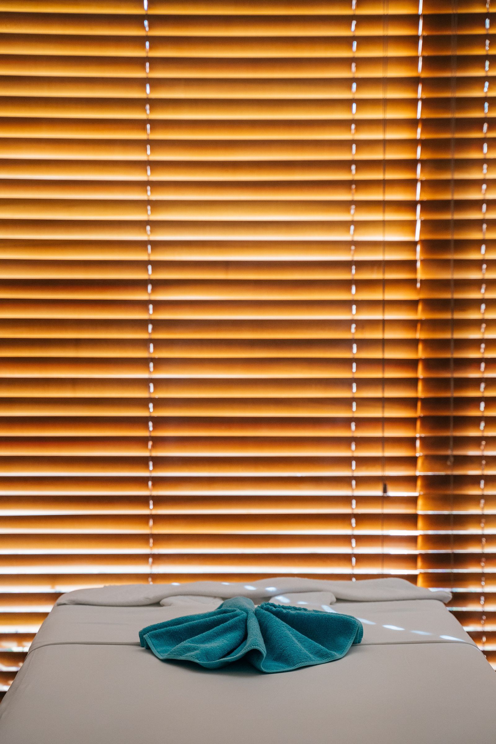 A treatment table dressed with a folded teal towel in front of wooden blinds