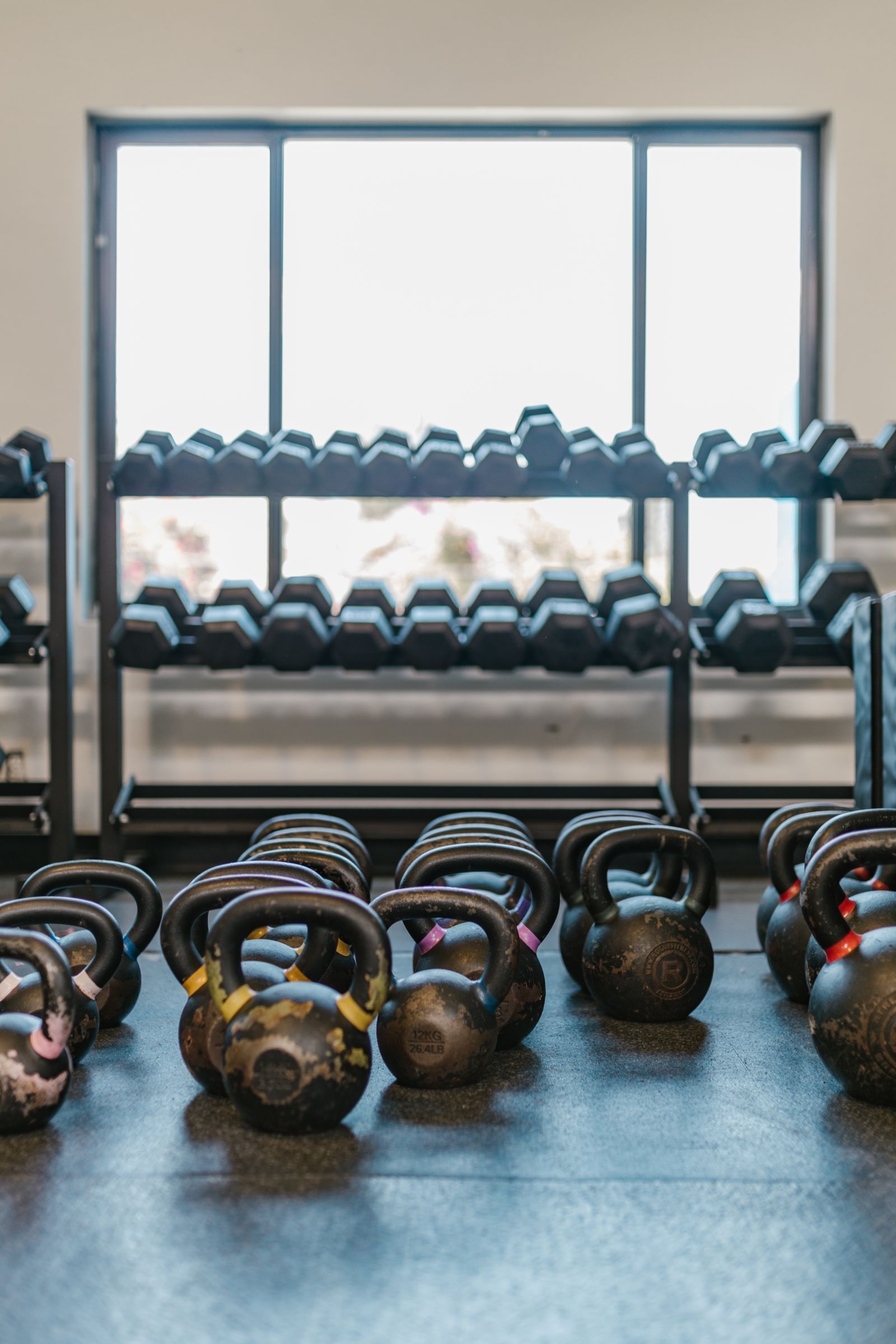 Kettlebells on the gym floor in front of dumbbell racks and a big bright window