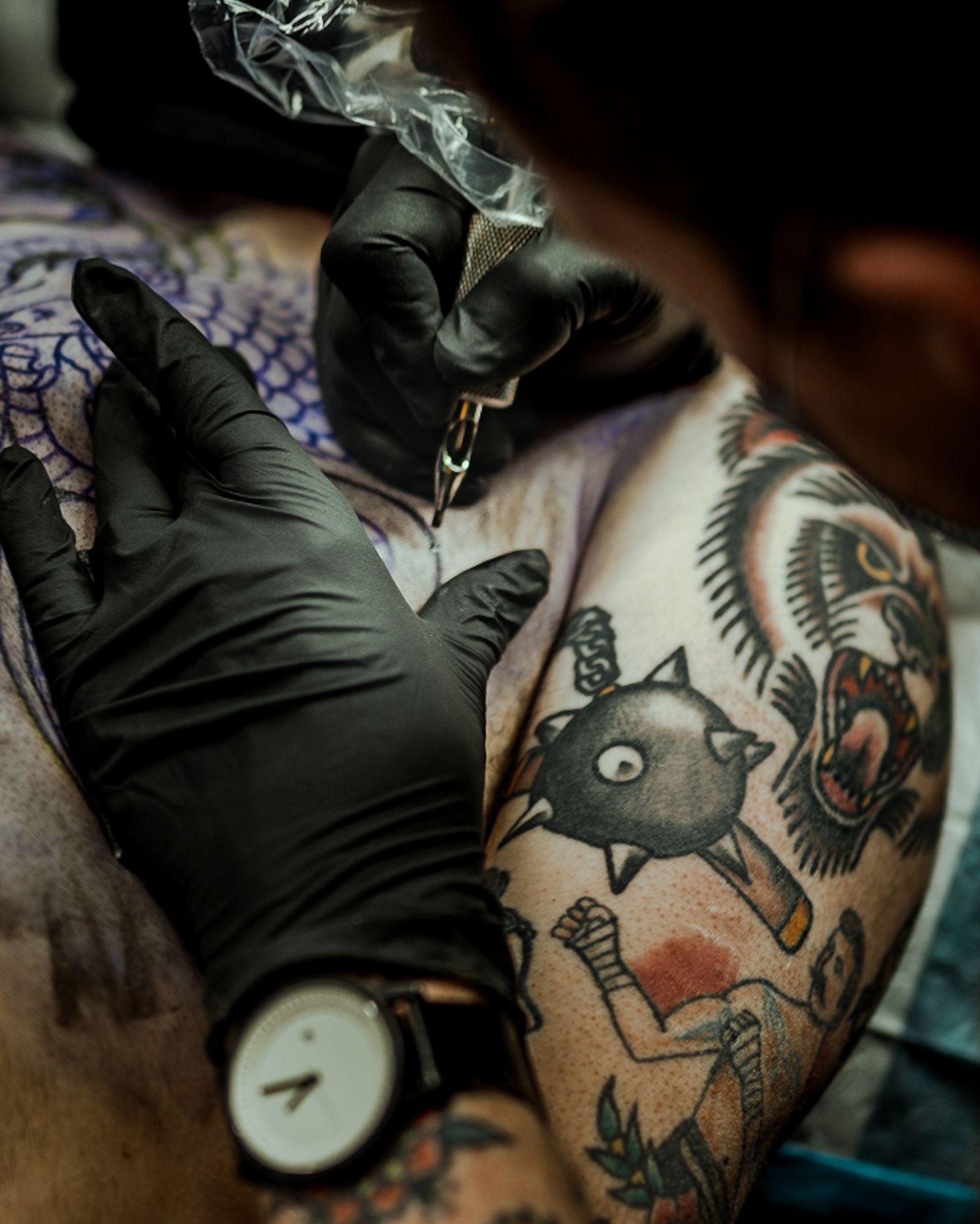 An artist in black gloves tattooing a client's shoulder covered in traditional work