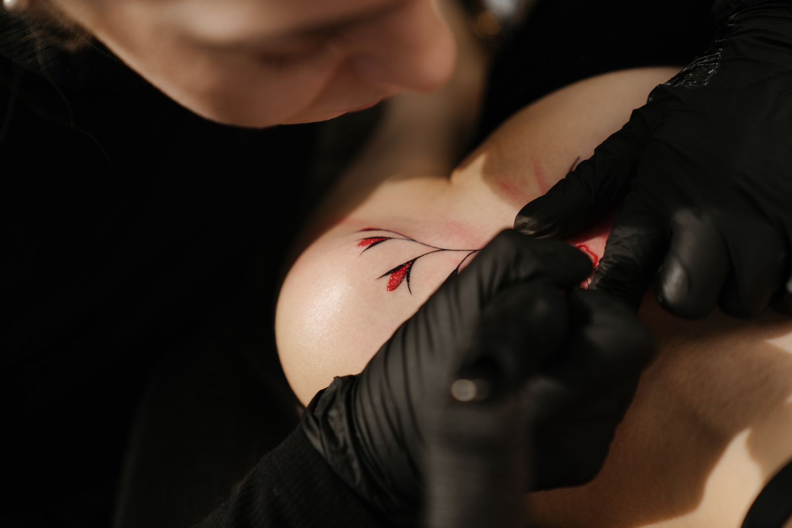 An artist in black gloves tattooing a fine red branch on a shoulder