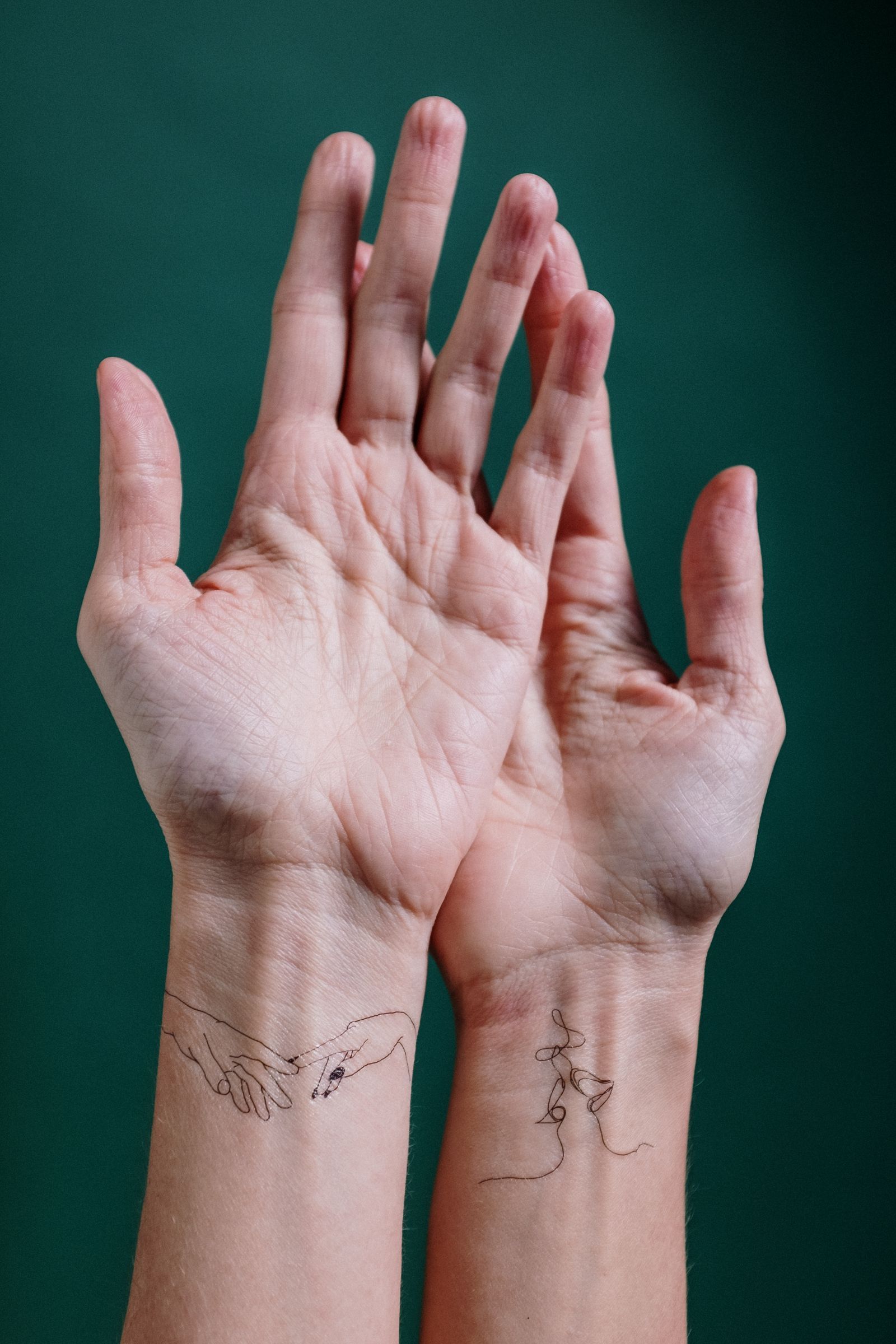 Two inner wrists with delicate single-line tattoos of hands and a face